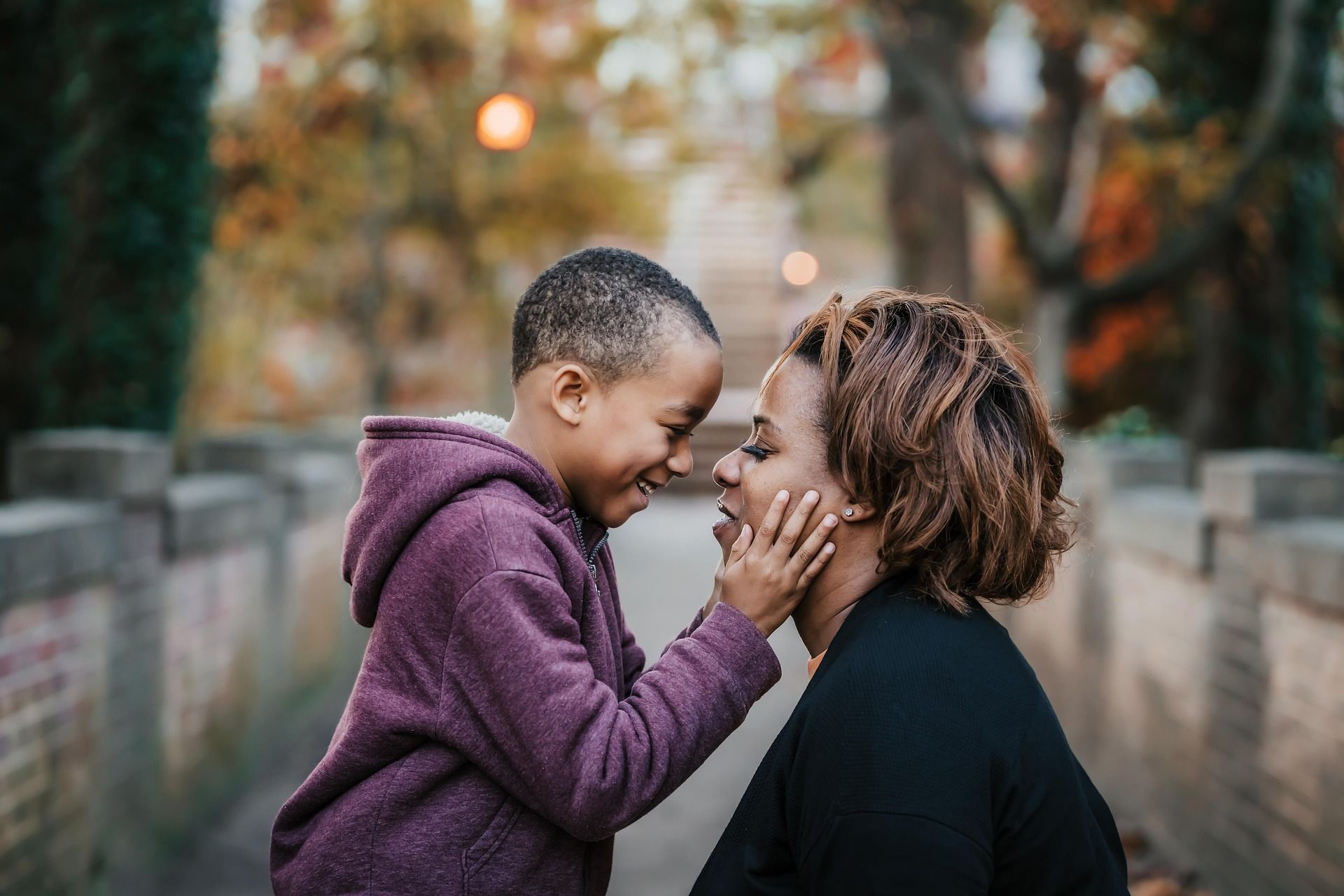 Boy touches mother's face, both smiling, outdoors on a stone walkway. Blurred fall foliage in background.