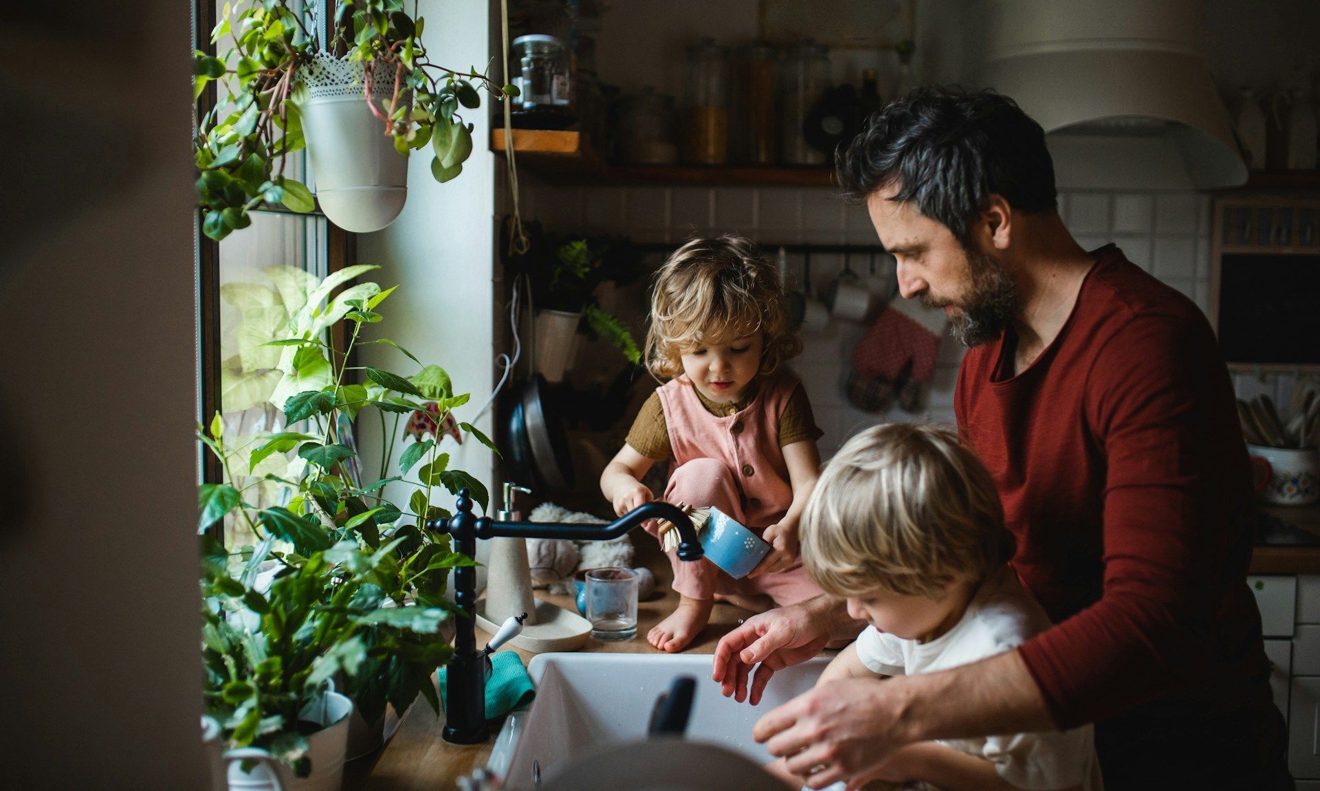Man helping two young children wash their hands at a kitchen sink near a window with plants.