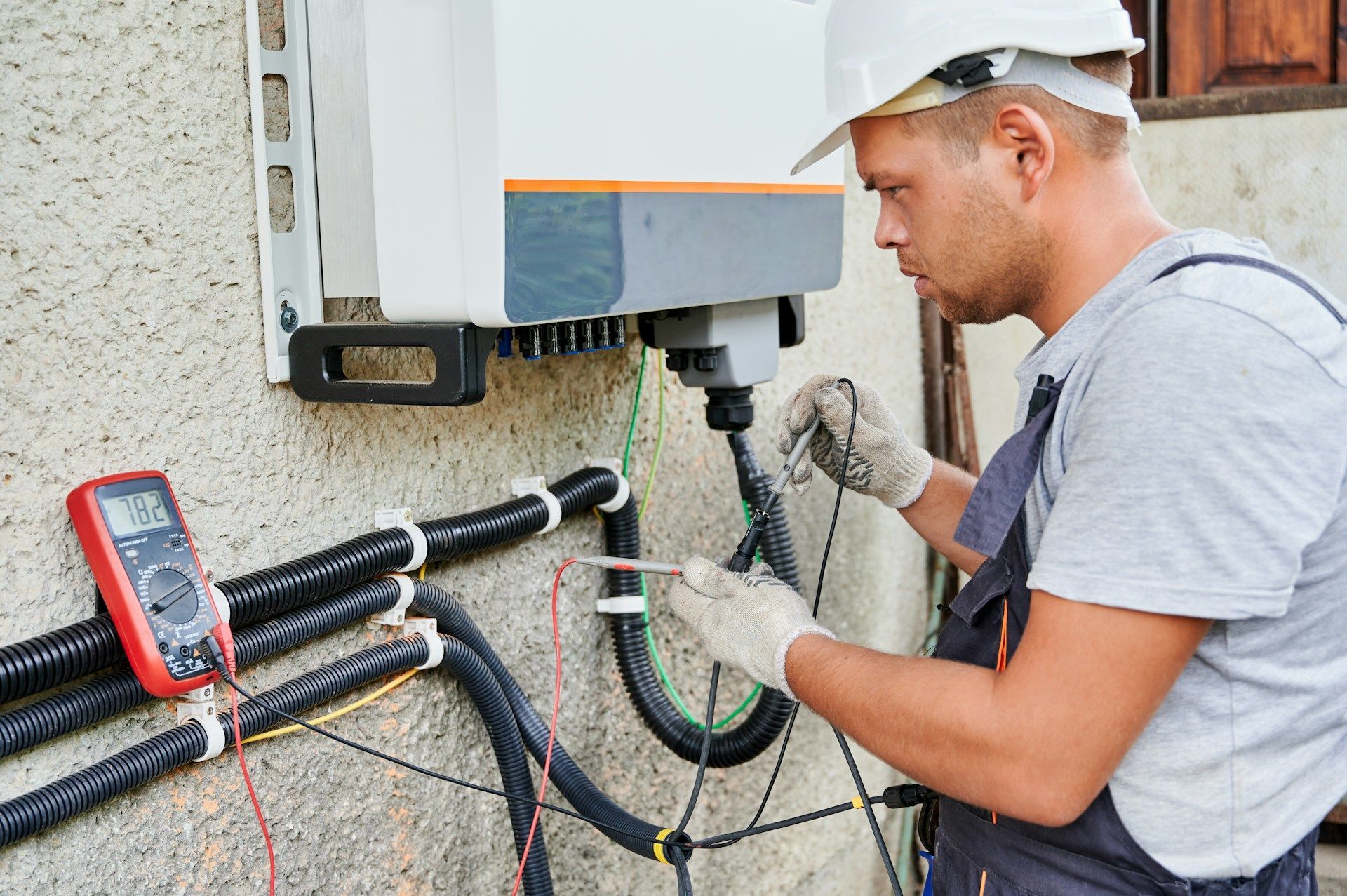 Electrician in a white helmet and gloves testing wires connected to a solar panel inverter.