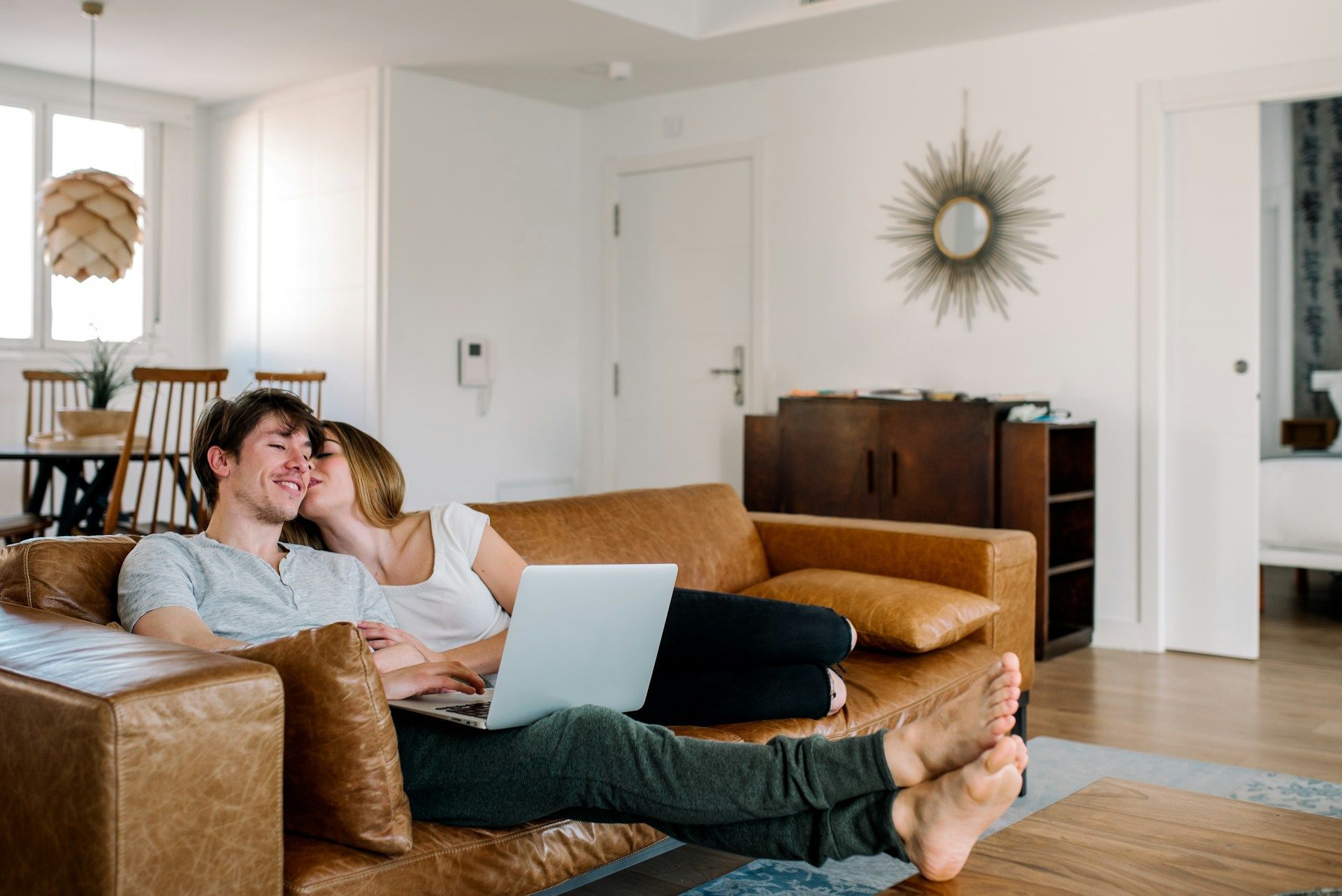 Couple relaxing on a brown sofa, one using a laptop, other kissing his cheek in a bright living room.