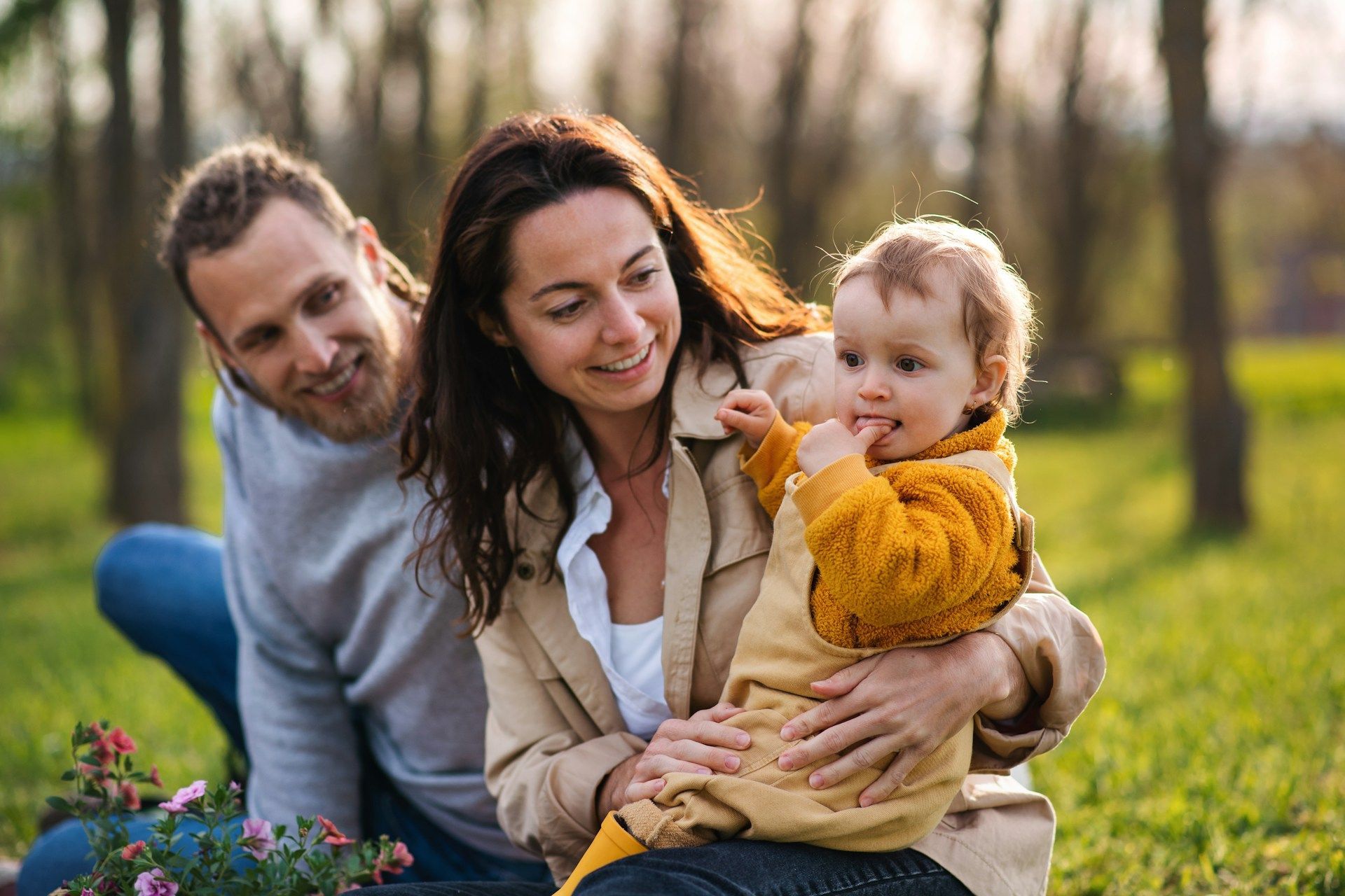 Family with baby in park, woman holding child, man smiling. Sunlight, green grass, trees.