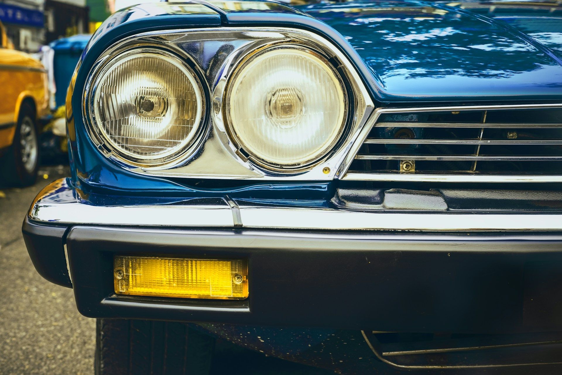 Close-up of a blue classic car's front with dual headlights, chrome details, and a yellow fog light.