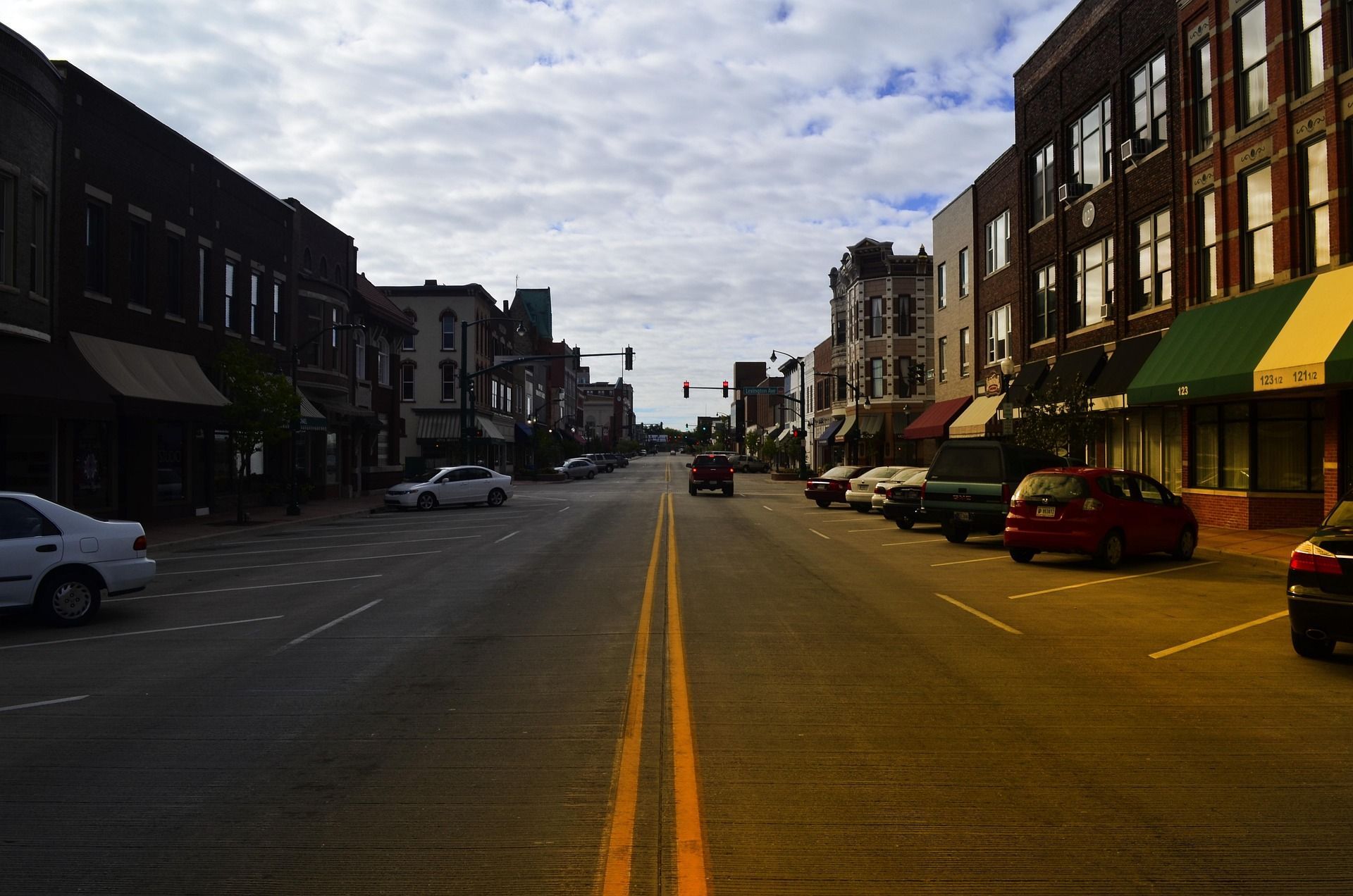 Street view of buildings and parked cars, under cloudy sky.