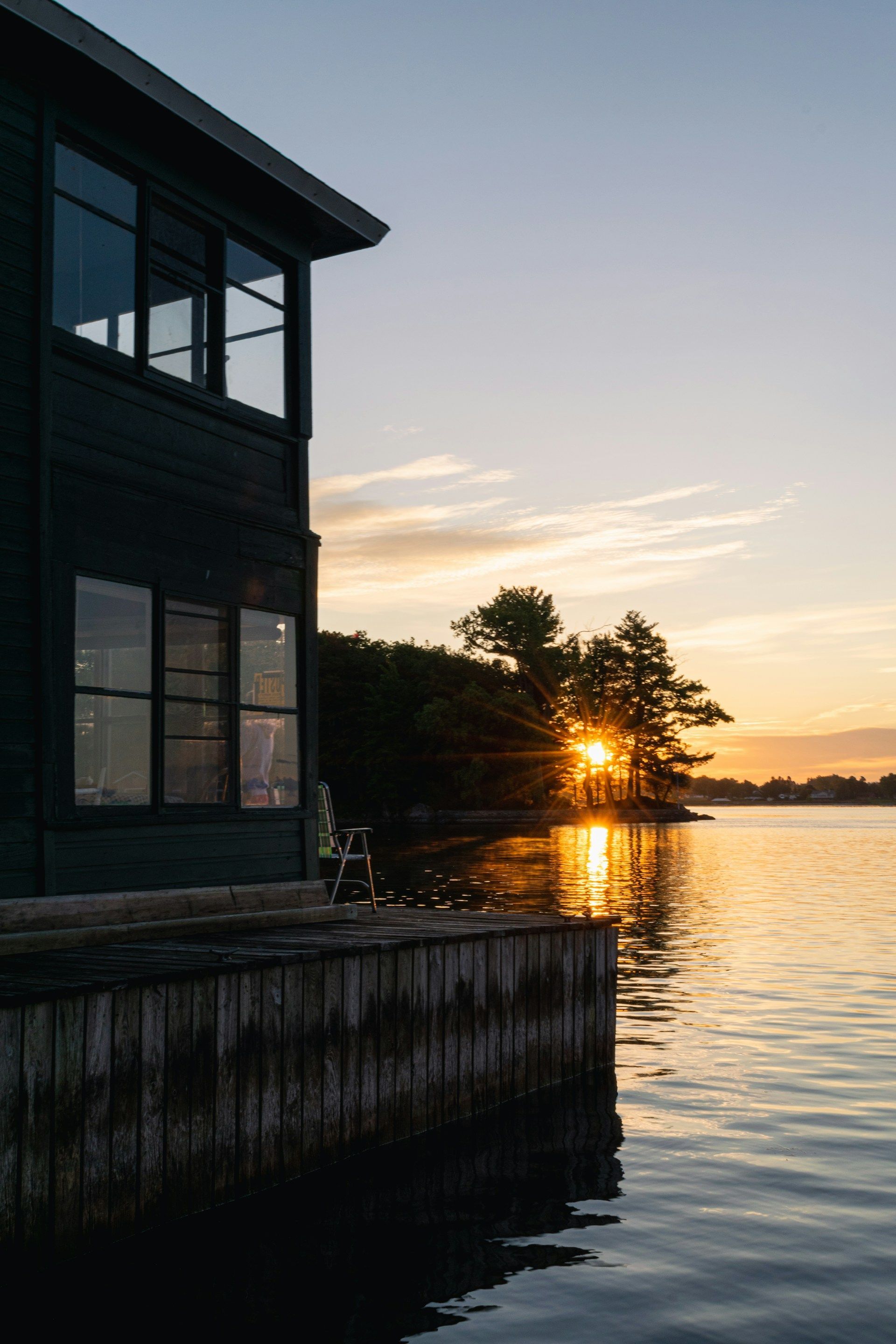 Dockside building at sunset, water reflecting golden light, sun peaking through trees on the horizon.