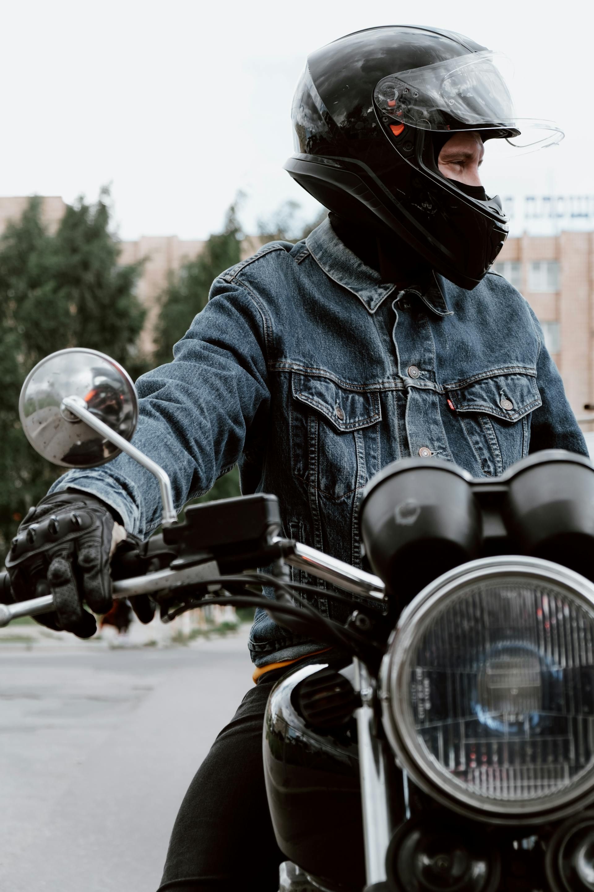 Person wearing a helmet and denim jacket on a motorcycle, holding the handlebars.