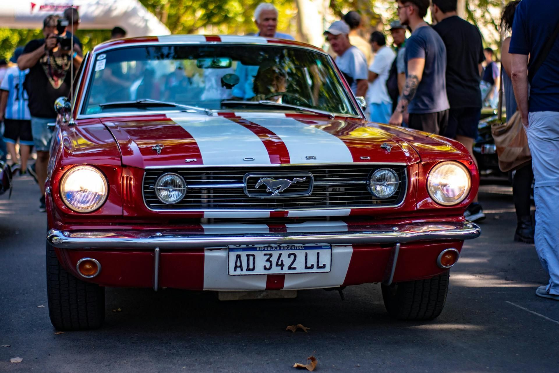 Red and white Ford Mustang with racing stripes, parked in street, surrounded by onlookers.