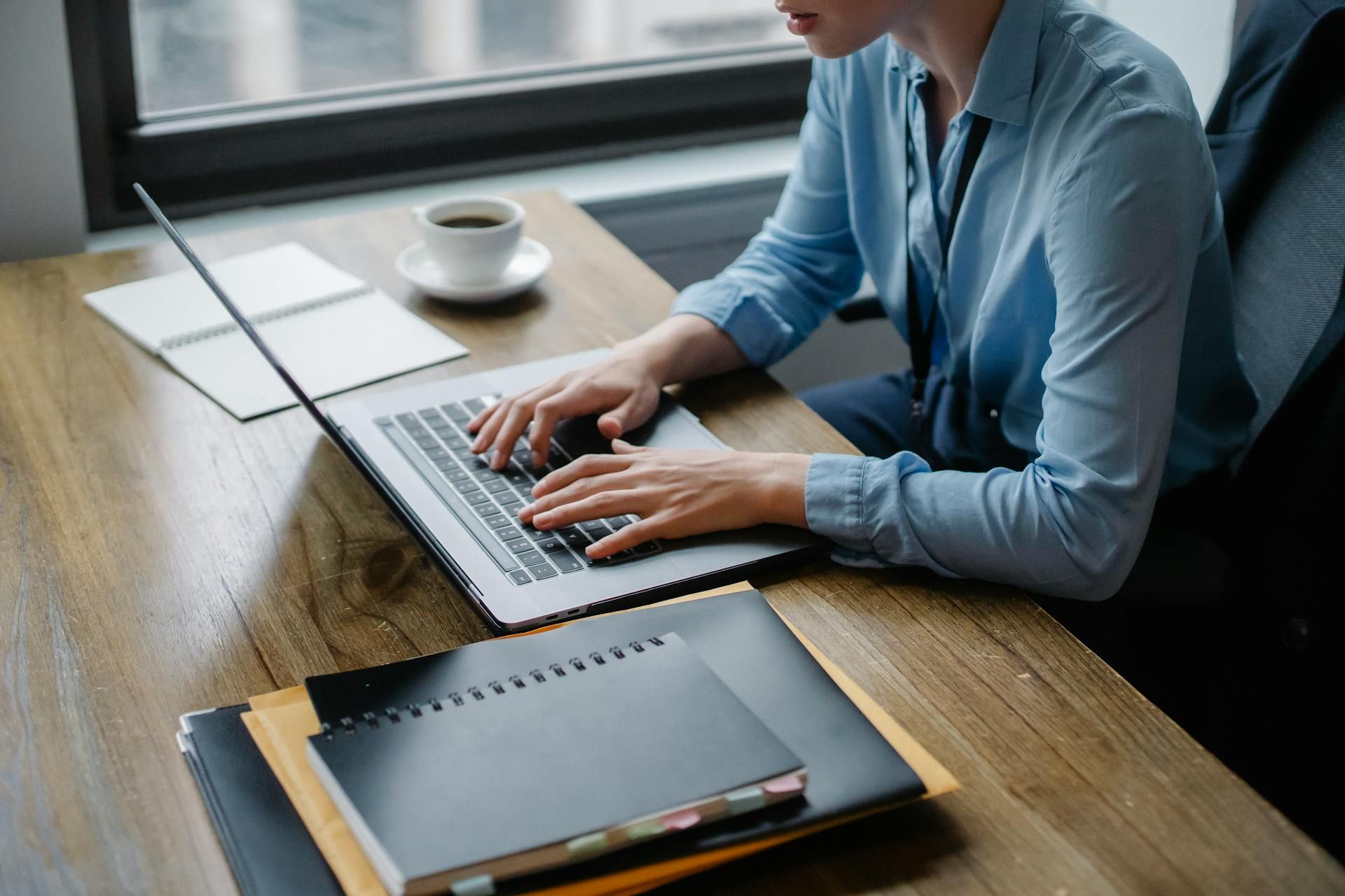Woman typing on laptop at a wooden desk with a coffee cup, notebook, and binders near a window.