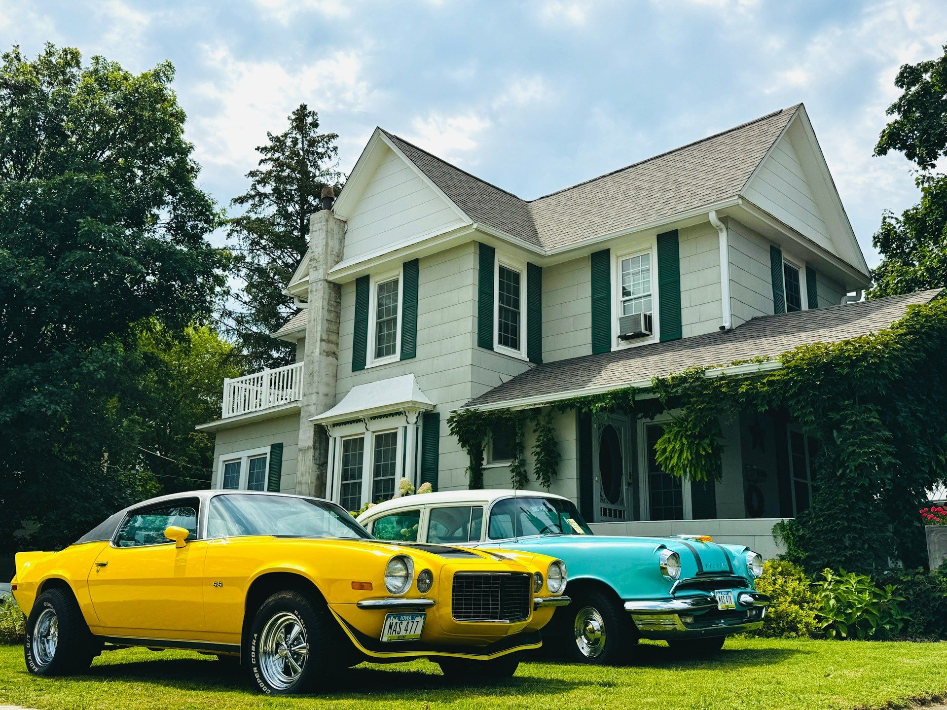 Yellow sports car and blue classic car parked on lawn in front of a light-colored house with green shutters.