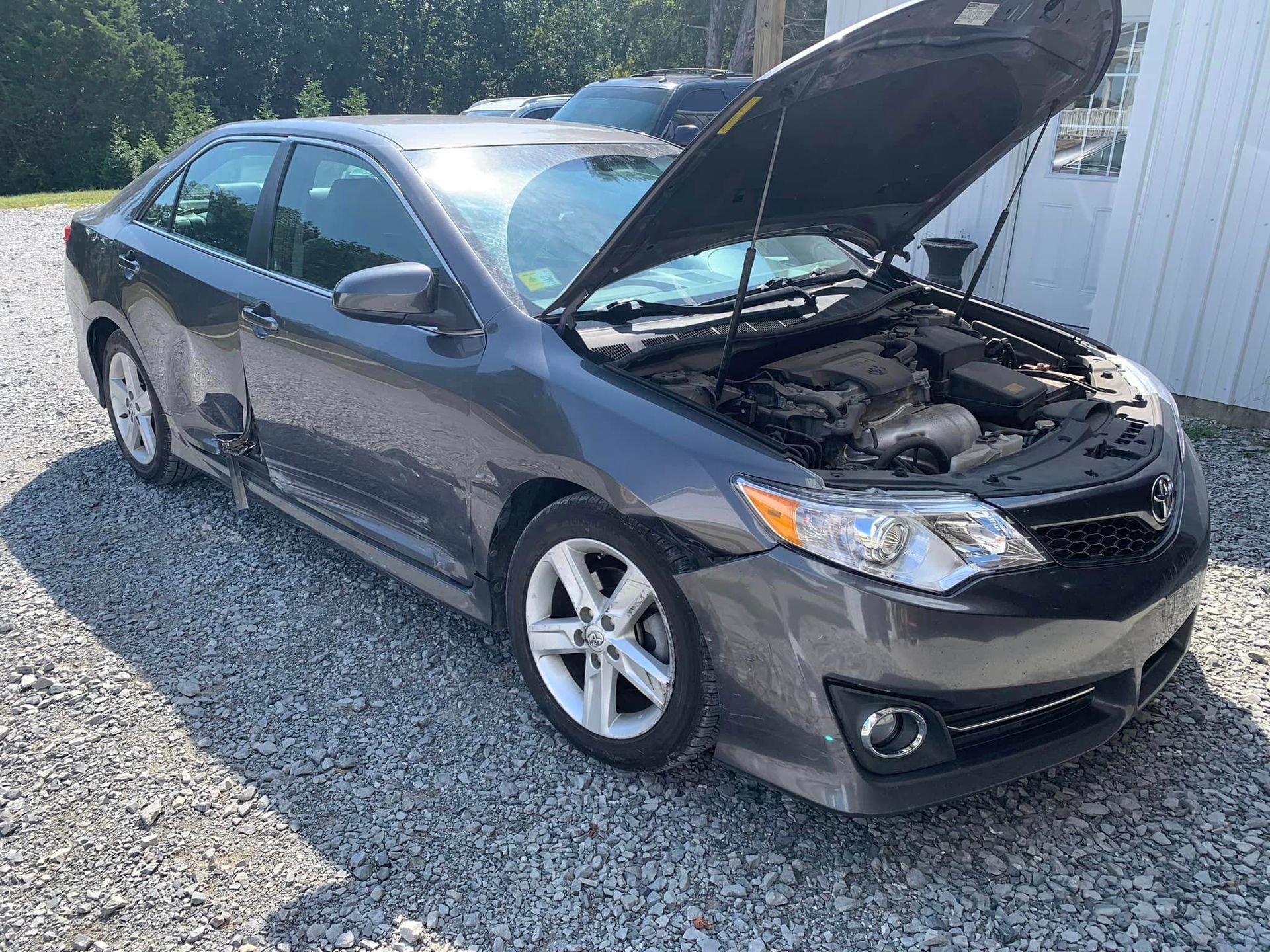 a gray car with the hood up is parked on gravel .
