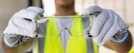 Persona con chaleco de seguridad sosteniendo gafas de seguridad transparentes. Lleva guantes blancos con puntos negros.