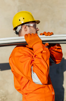 Obrero de la construcción que lleva tubos blancos sobre el hombro, vistiendo casco amarillo y ropa de trabajo naranja.