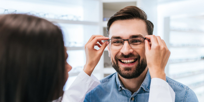 In einem hellen Optikergeschäft hilft ein Optiker einem lächelnden Patienten beim Anprobieren einer Brille.
