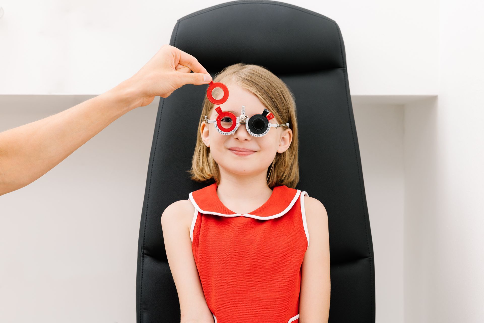 Während einer Augenuntersuchung trägt ein Kind eine Probebrille mit einer roten Linse über einem Auge, während eine Hand eine Ersatzlinse hält.