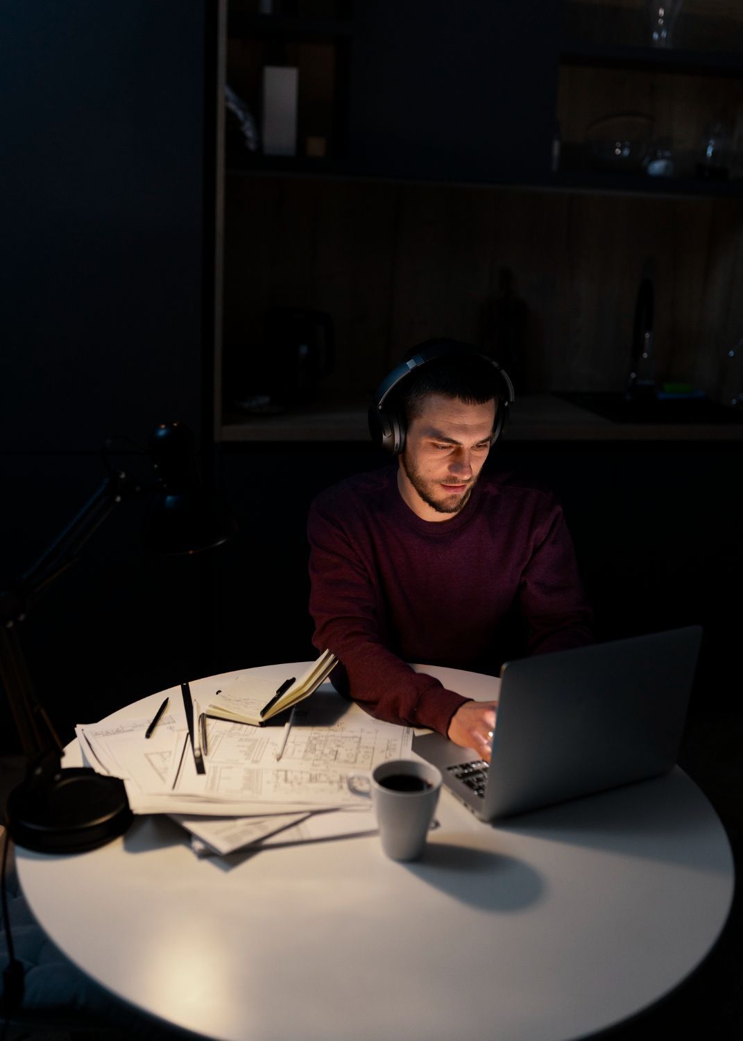 Un hombre con auriculares trabaja con una laptop en una mesa redonda en una habitación oscura. Hay documentos, bolígrafos y una taza de café dispersos.