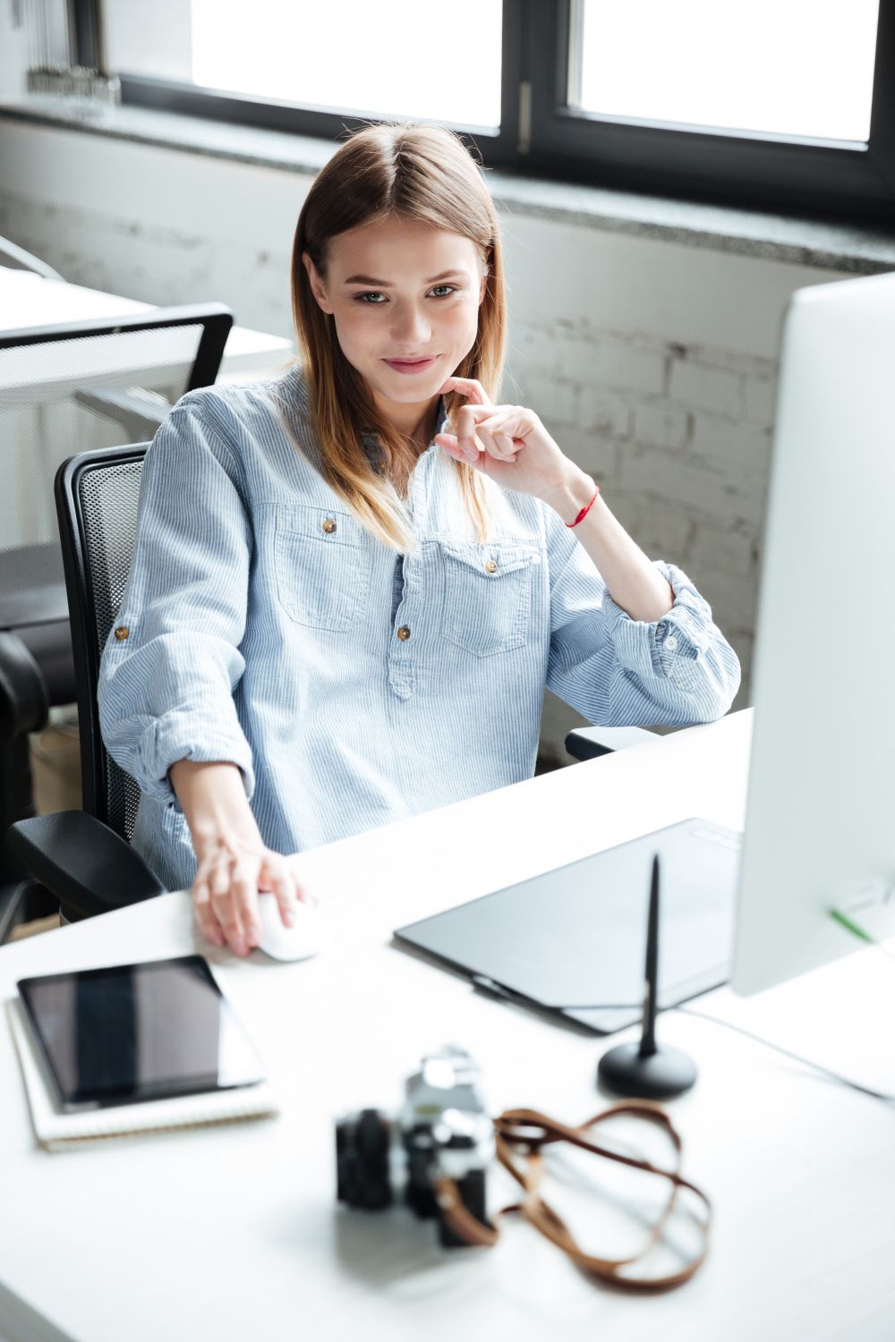 Una mujer con camisa azul trabaja en un escritorio con una computadora, un ratón, una tableta y una cámara. Sonríe y hay una ventana al fondo.