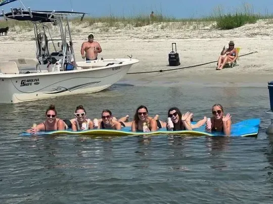 Seven people on a floating mat in the water, boat and beach in background. Sunny day.