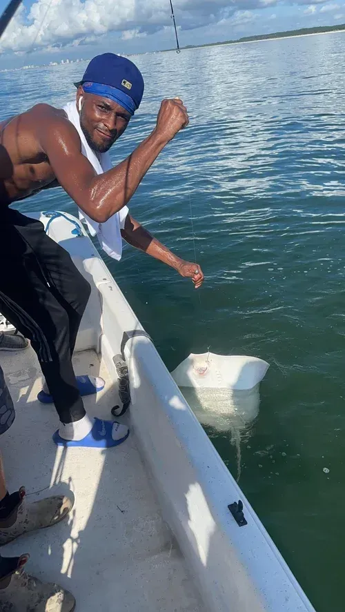 Man on boat, reeling in a large, white stingray. Blue water, clear sky. He is shirtless, smiling, fist raised.