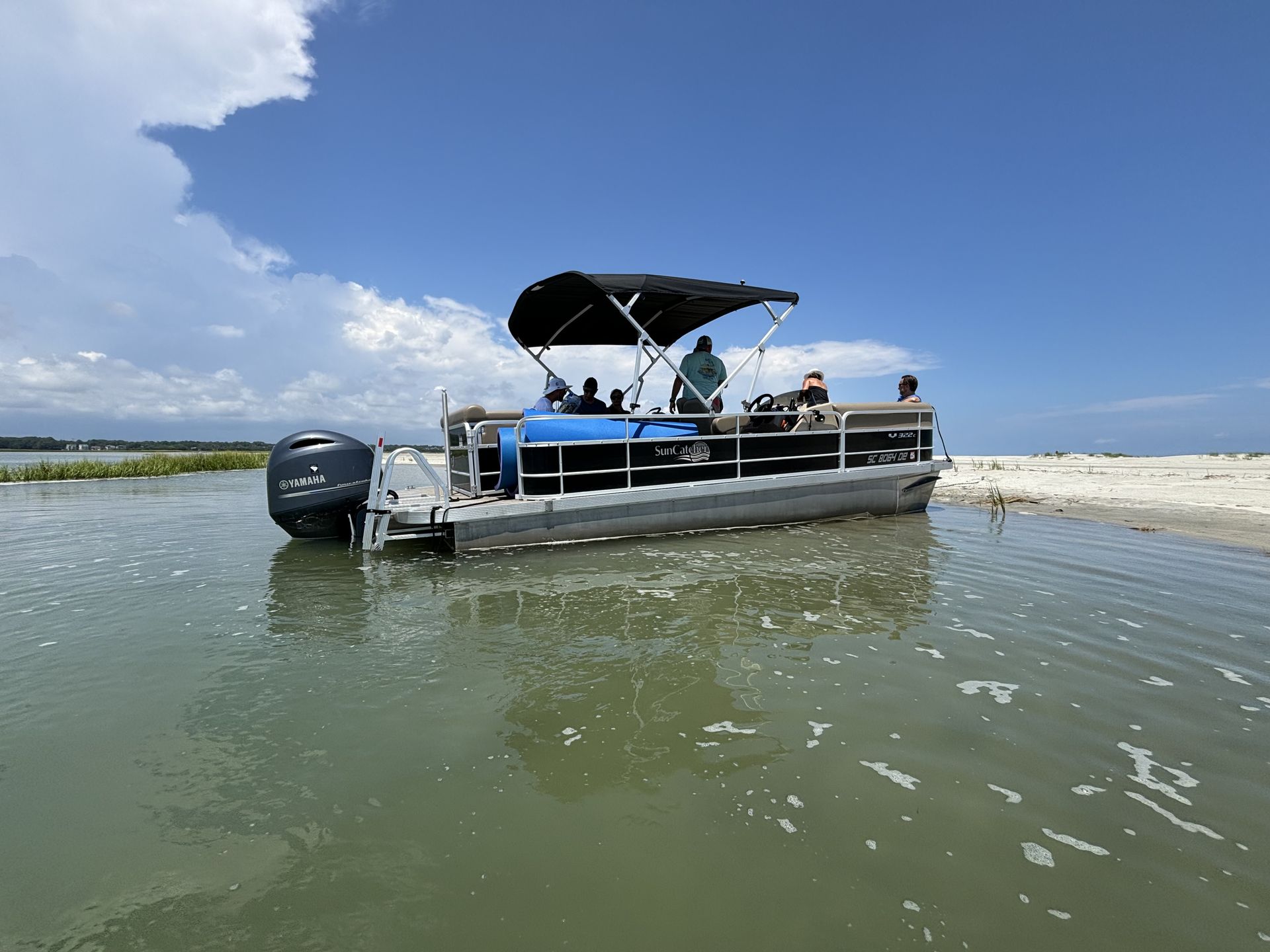 Pontoon boat cruising on dark water under a blue sky.  Trees line the shore.