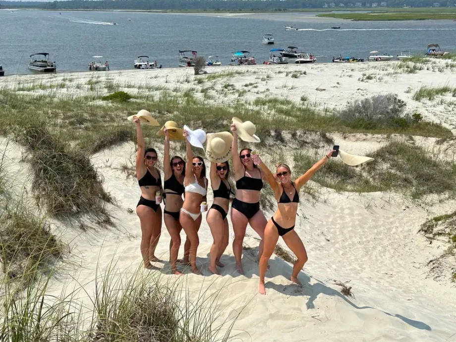 Group of women in bikinis on a sandy dune, raising hats towards boats in the water.