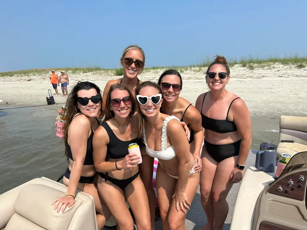 Six women in swimsuits smile on a boat near a beach; sunny day, blue sky.