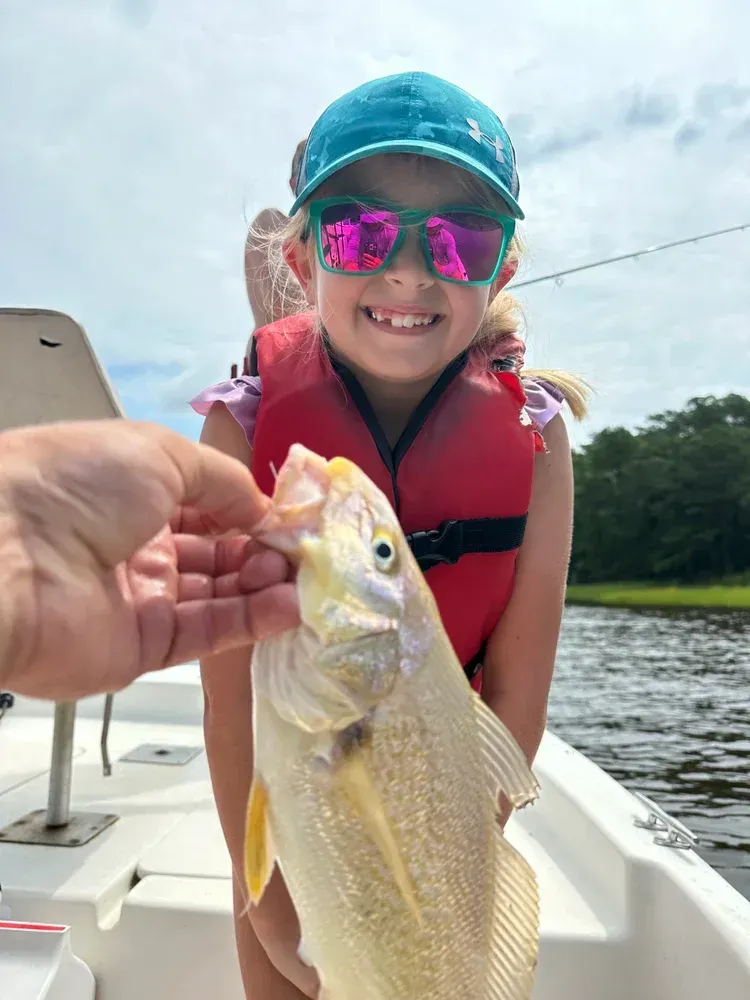 Young person in a life vest smiles, holding a fish. Boat on water, sunny day.