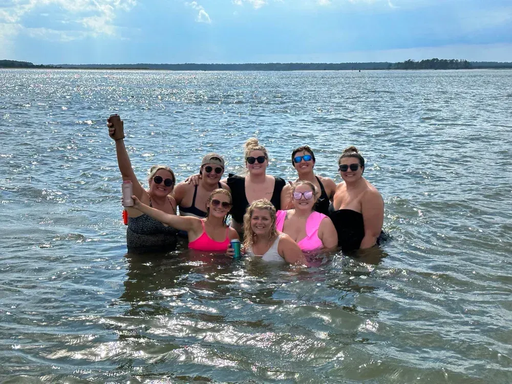 Group of women standing in a body of water, some wearing sunglasses, with sunny sky in background.