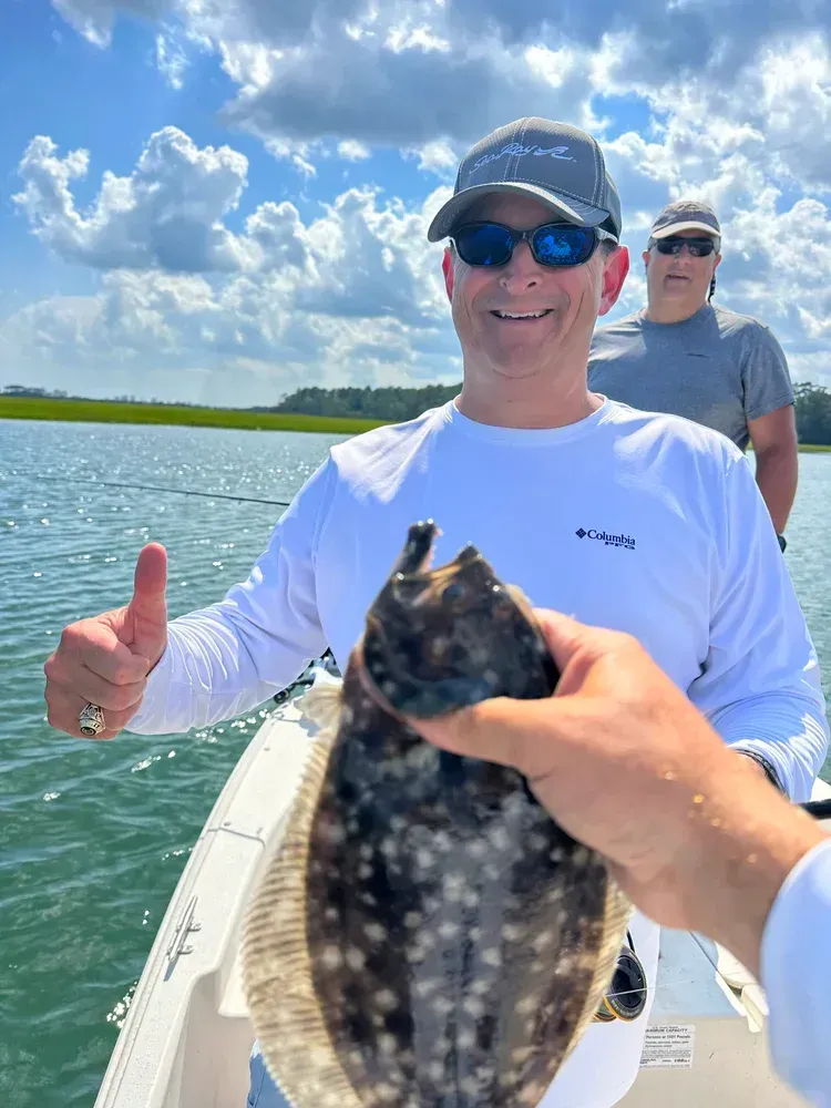 Man on boat, smiling and giving thumbs up, holding a speckled fish; blue sky and water.