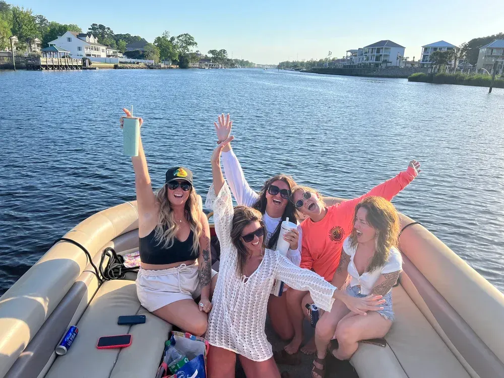 Group of people on a boat, waving and smiling. River setting with buildings and trees in the background. Sunny day.