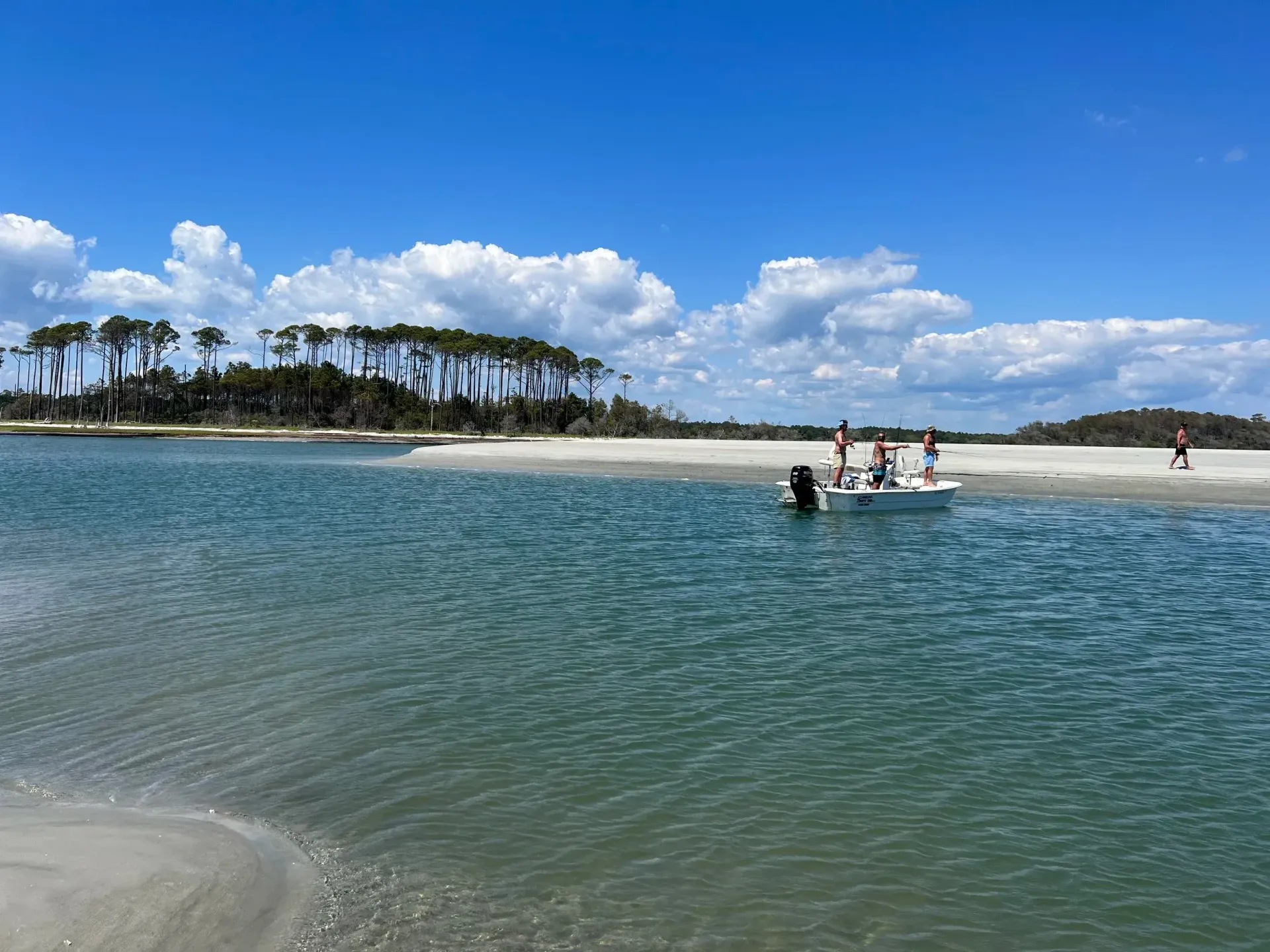 Coastal scene: Calm blue water, sandy beach, small boat, people, palm trees, blue sky with clouds.