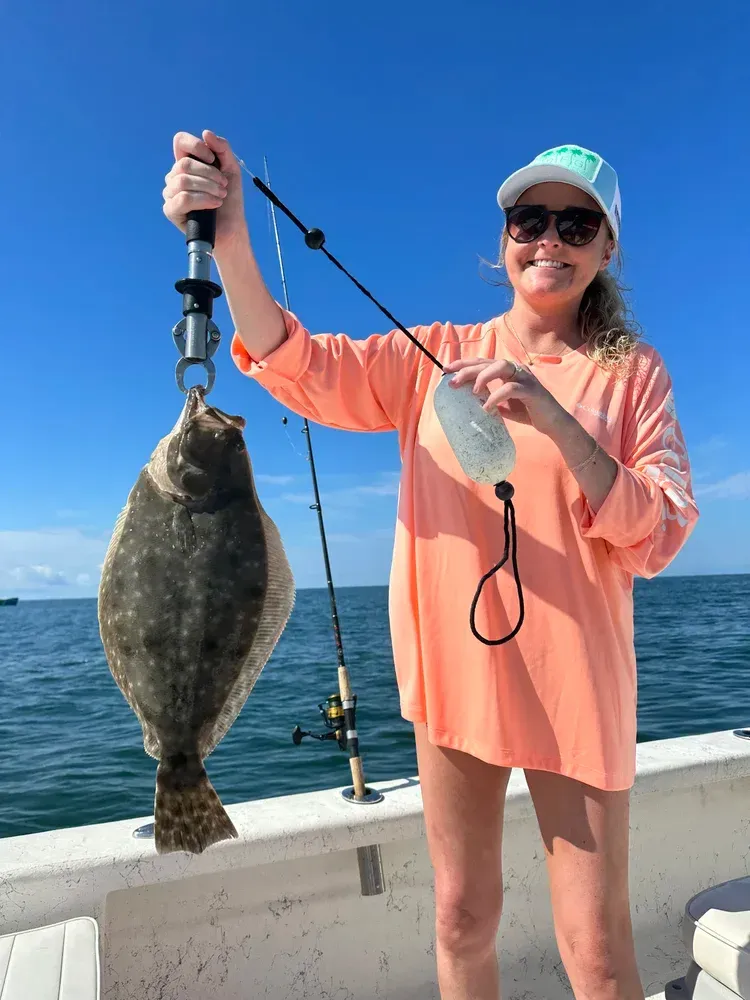 Woman holding a large flounder caught on a boat, wearing sunglasses and an orange shirt, blue sky in the background.