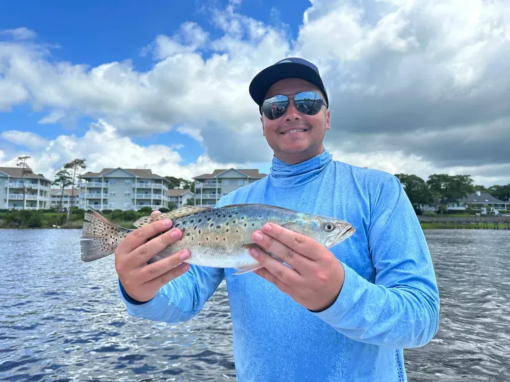 Man holding a speckled trout, smiling, blue shirt, sunglasses, on a boat with buildings in the background.