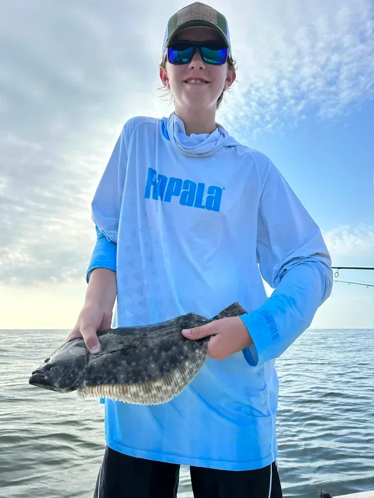 Young person smiles holding a speckled flounder on a boat under a cloudy sky.