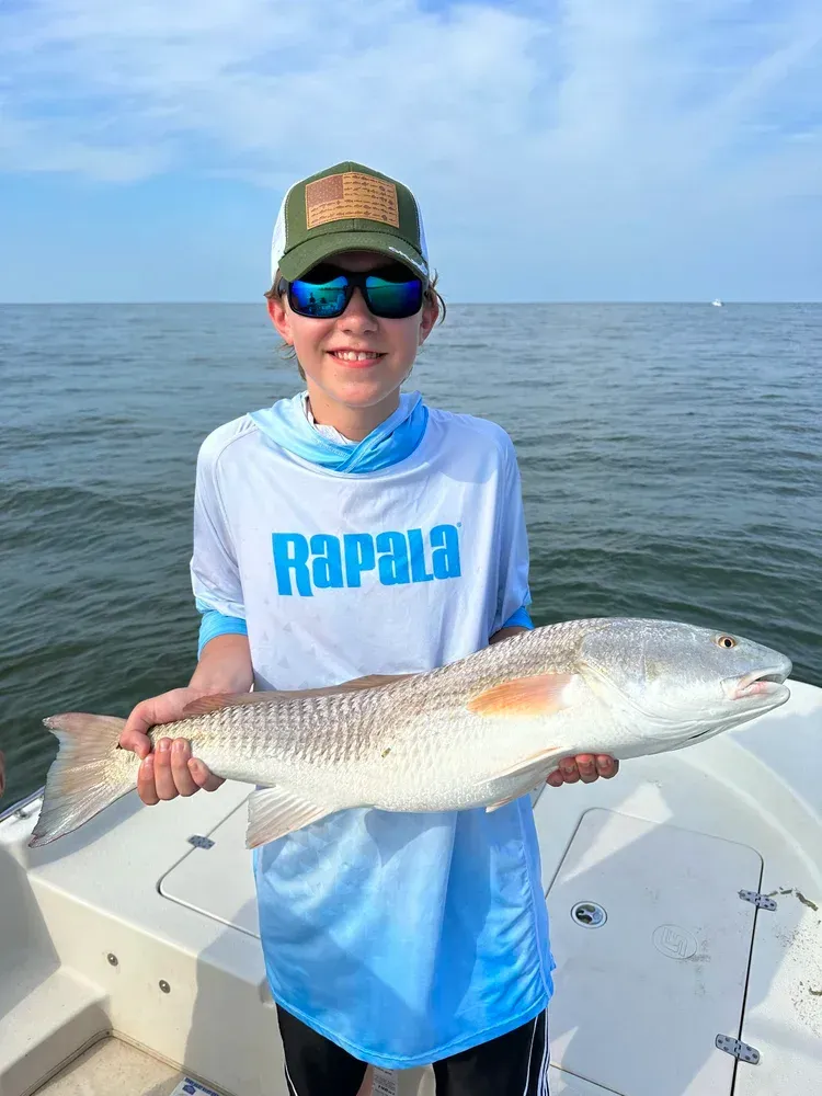 Boy on a boat smiles, holding a large redfish. Wearing hat, sunglasses, Rapala shirt, and fishing.