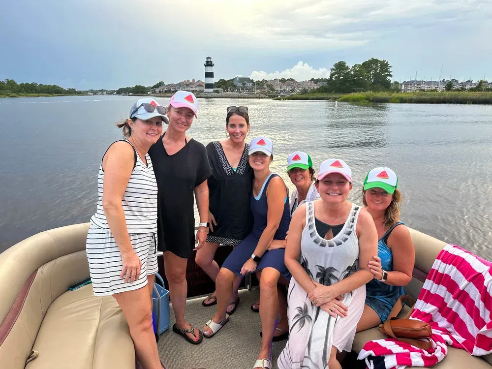 Group of women on a boat wearing matching hats, posing near a lighthouse.