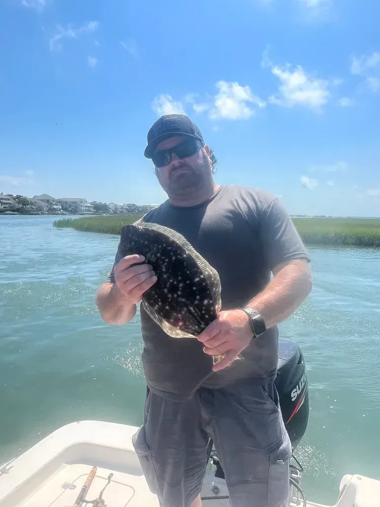Man on boat holds up a speckled flatfish, blue water, green marsh in background, sunny sky.
