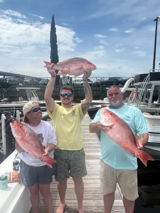 Three people holding up large red snapper fish at a dock, bright sky.