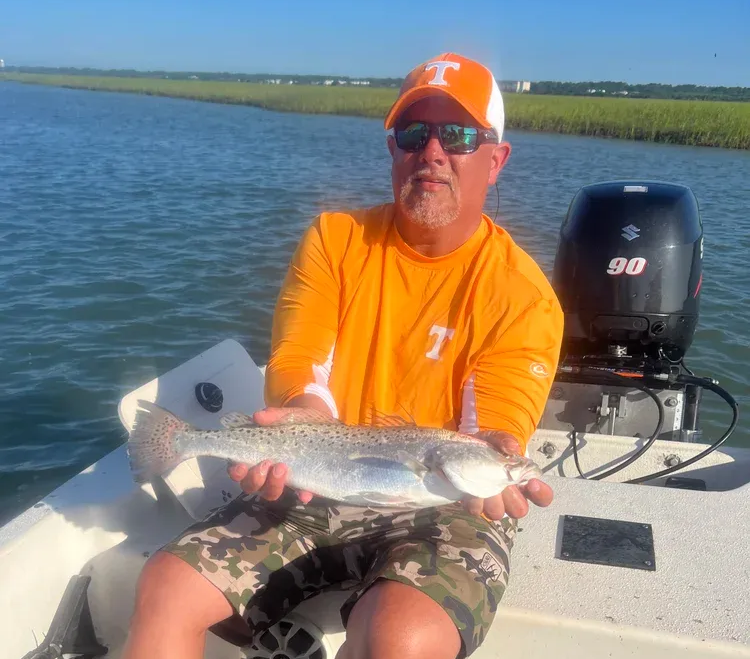 Man on a boat holding a speckled trout. He wears an orange shirt and cap. A motor is in the background.