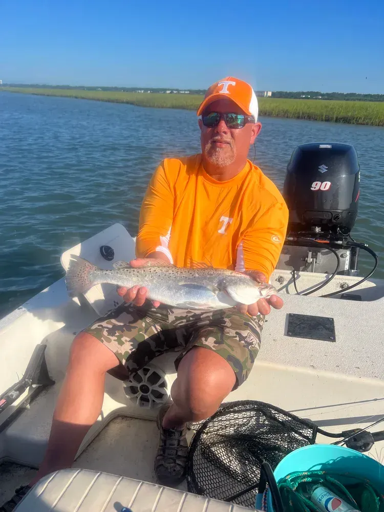 Man in orange shirt holding a speckled trout on a boat in a marsh, under a sunny sky.