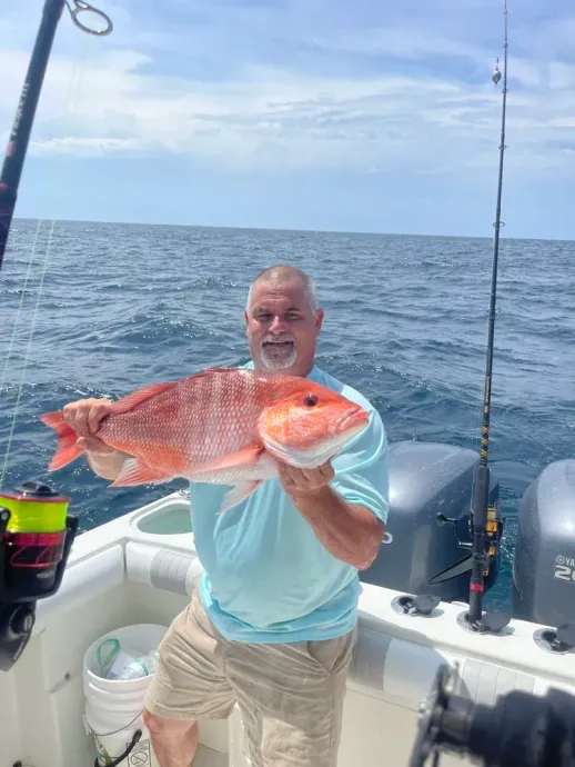 Man on boat holding a large red snapper he caught. Ocean setting, sunny day, smiling.