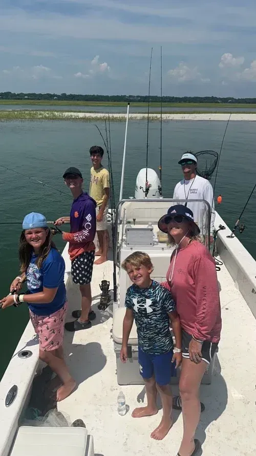 People fishing on a boat in a coastal setting. They are smiling. Cloudy sky and shoreline in the background.