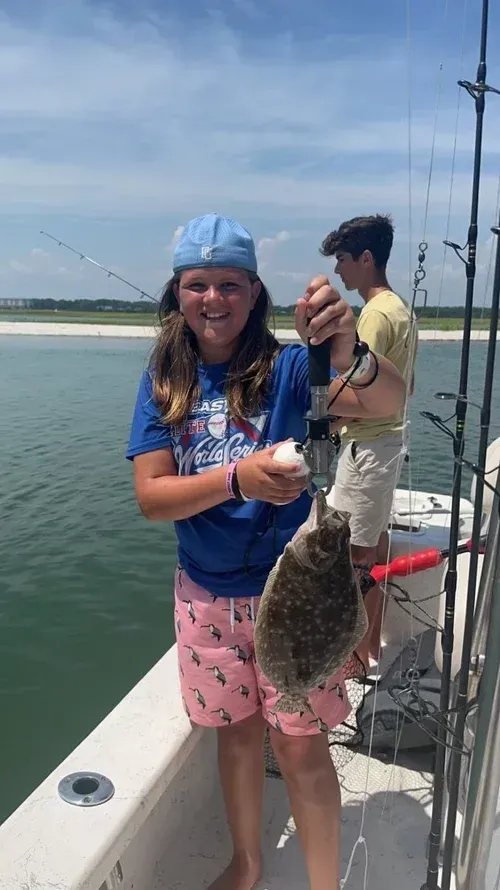 Young person on a boat holding up a fish, smiling. Another person in the background. Sunny day, water.