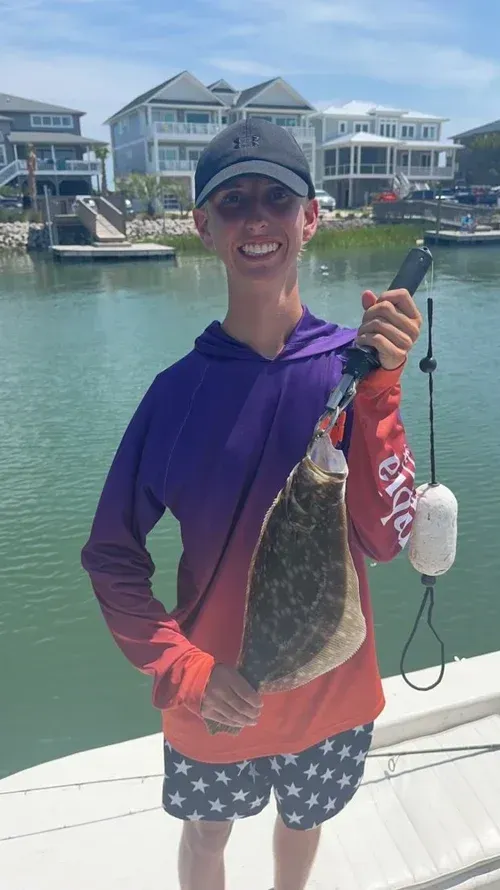 Young person smiling, holding a flounder caught while fishing. Wearing a hat and colorful shirt, standing near water.