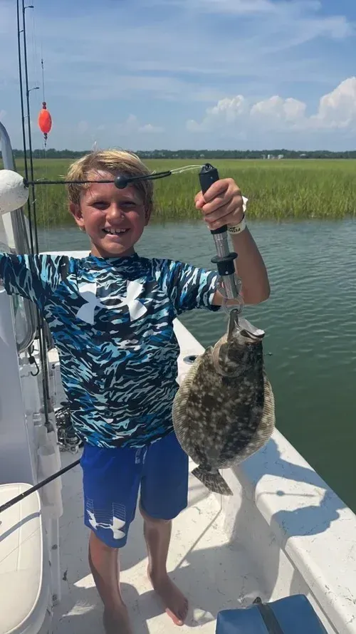 Boy smiles, holding a flounder on a boat; sunny day, water and marsh in background.