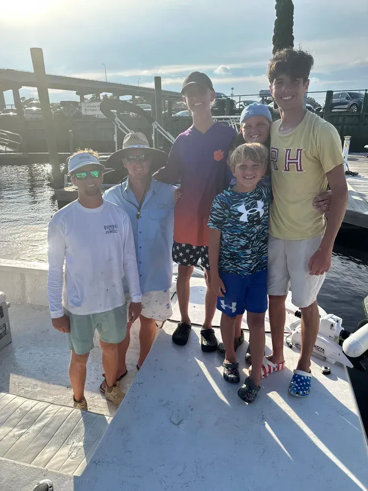 Group of people on a boat dock, smiling. Several wearing hats, shorts, and light-colored shirts on a sunny day.