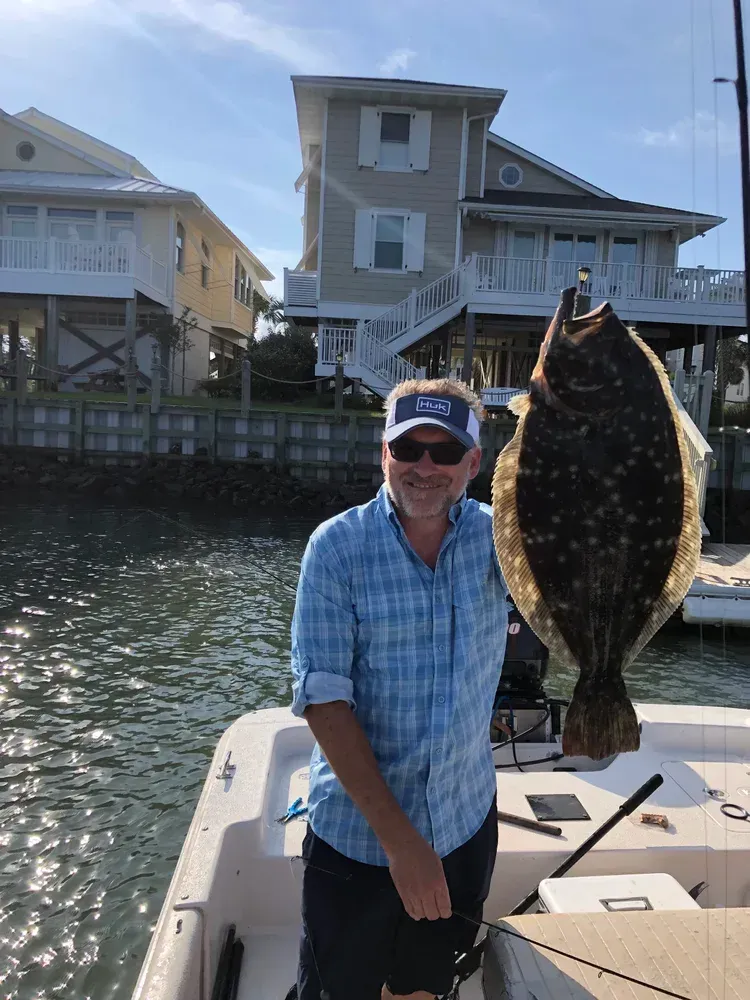Man holding large flounder on a boat, houses in the background. Sunny day.