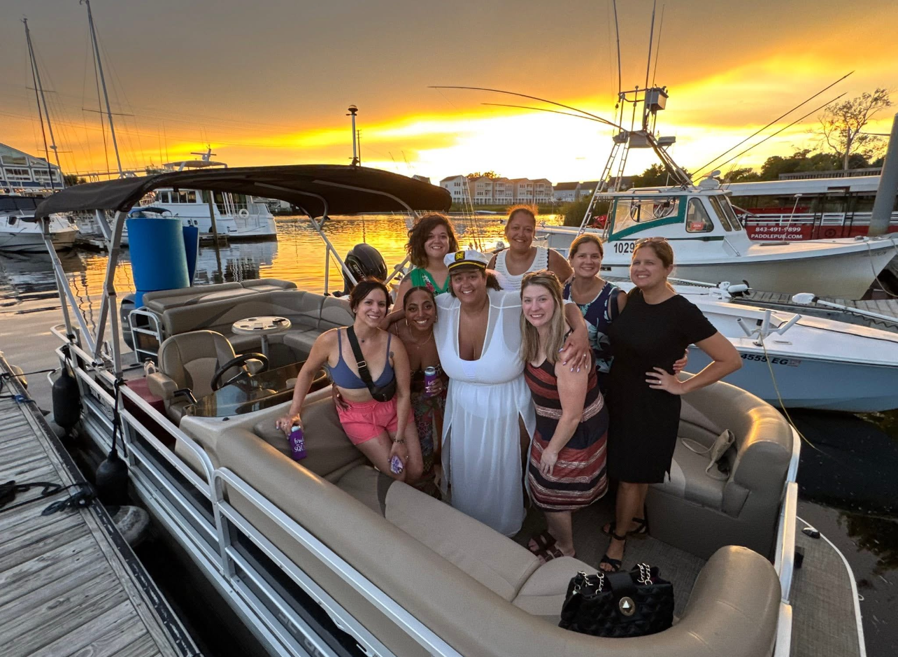Pontoon boat cruising on dark water under a blue sky.  Trees line the shore.