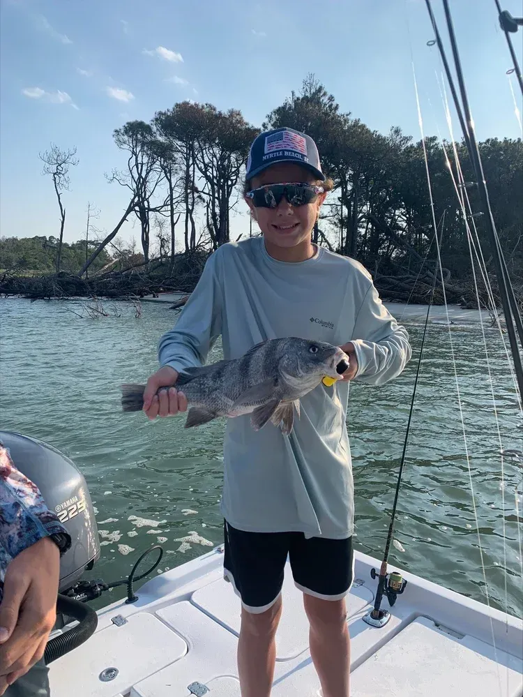 Boy on a boat smiles, holding a gray fish. Water and trees in the background. Sunny day.