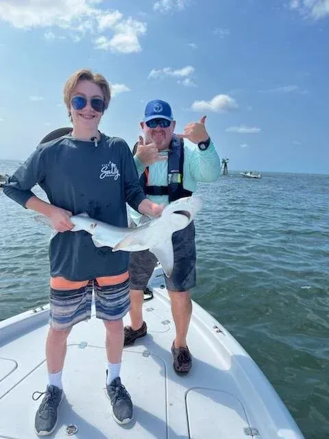 Two people on a boat, holding a small shark. One gives a thumbs-up. Bright sunny day, water visible.
