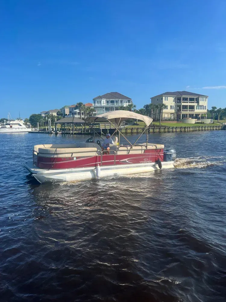 Pontoon boat on dark water with a red stripe, a canopy, and houses in the background on a sunny day.