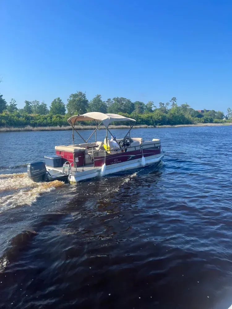 Pontoon boat on dark water with outboard motor. Sunny day, trees in background.